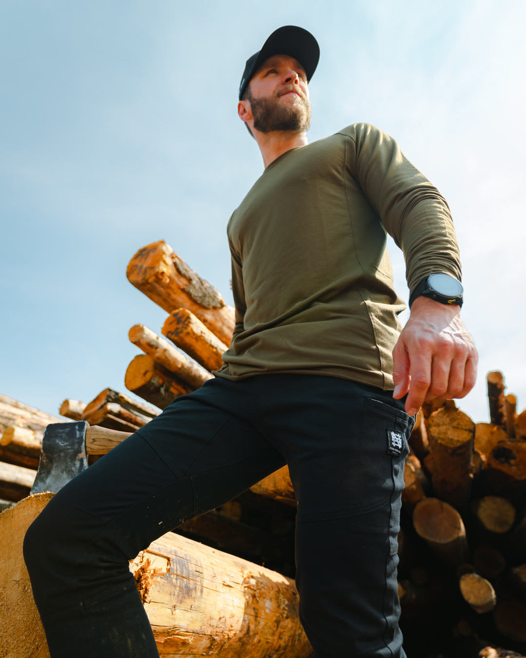 Man in a green shirt and black pants standing among stacked logs with a clear blue sky.