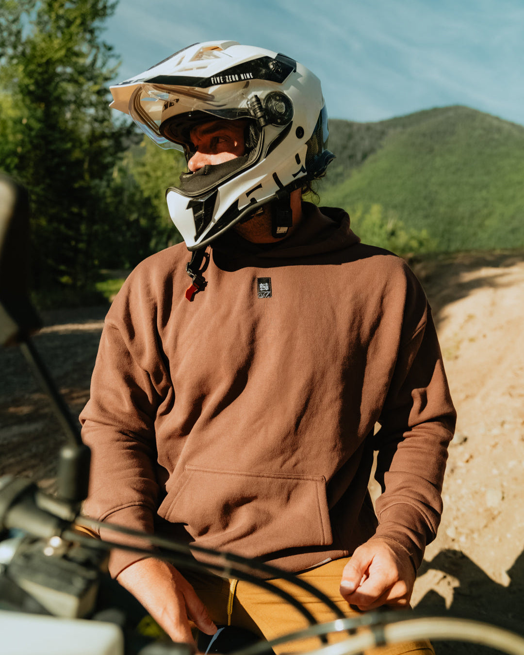 Person wearing a helmet and brown hoodie sitting on a motorcycle with mountains in the background