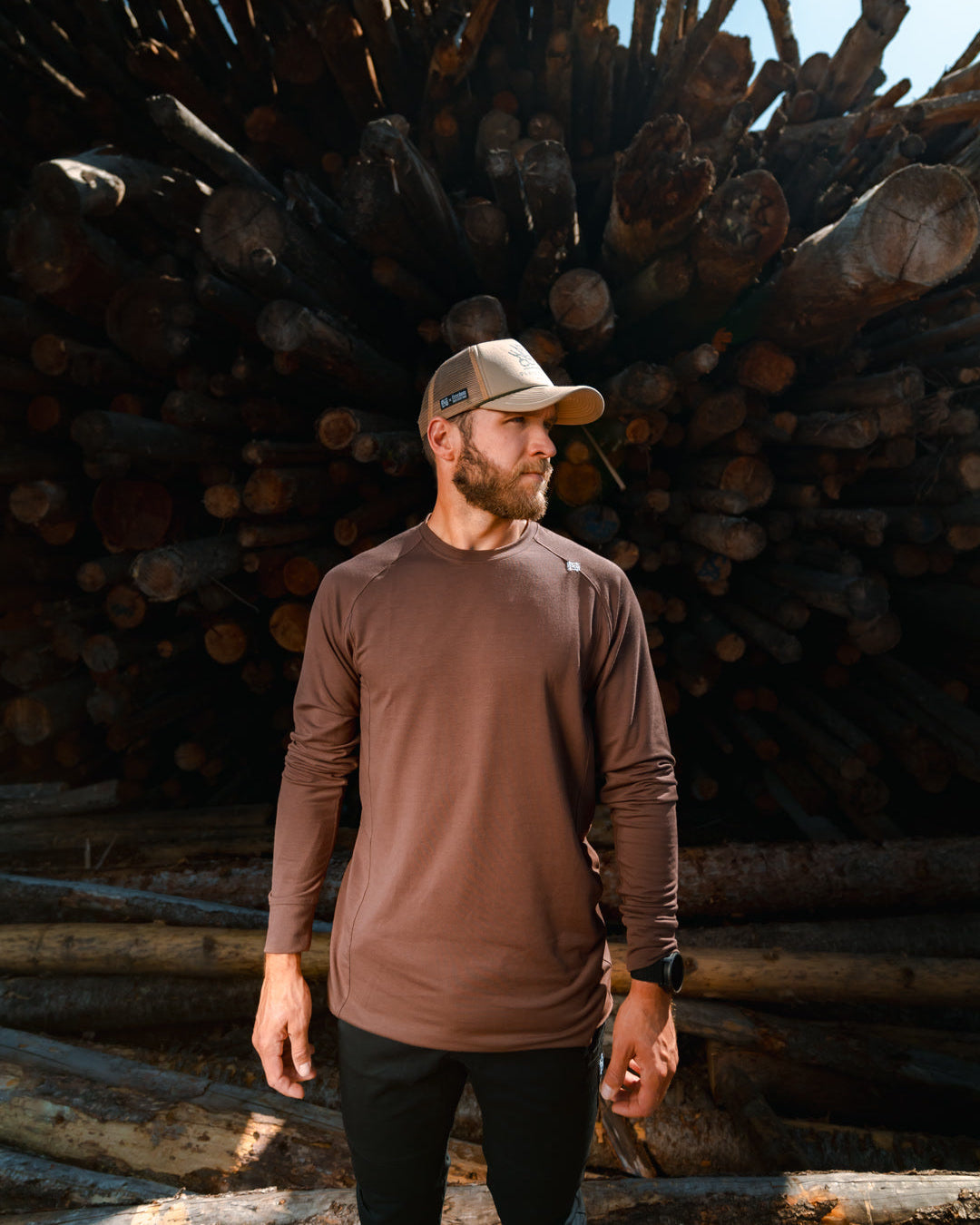 Man standing in front of a stack of logs wearing a brown long-sleeve shirt and cap.