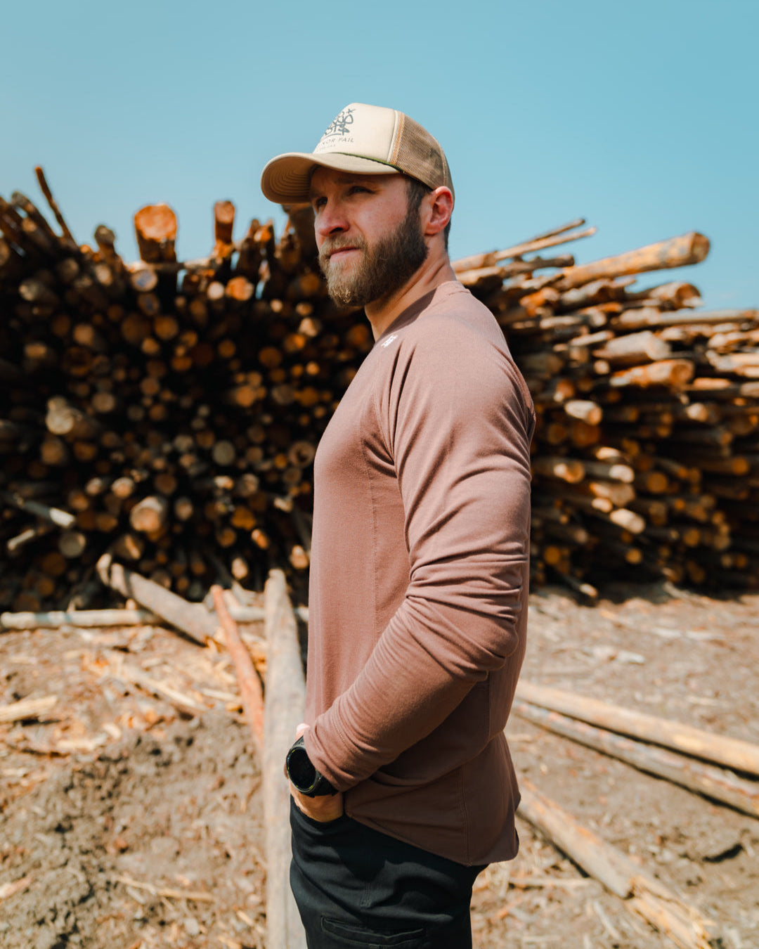 Man standing in front of a stack of logs with a clear blue sky.