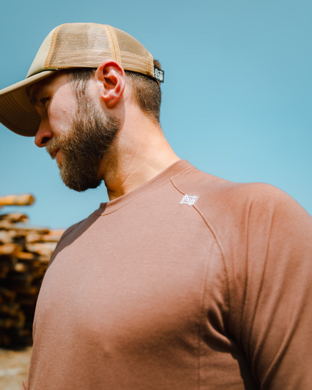 Man wearing a brown cap and shirt with a logo, standing against a blue sky.