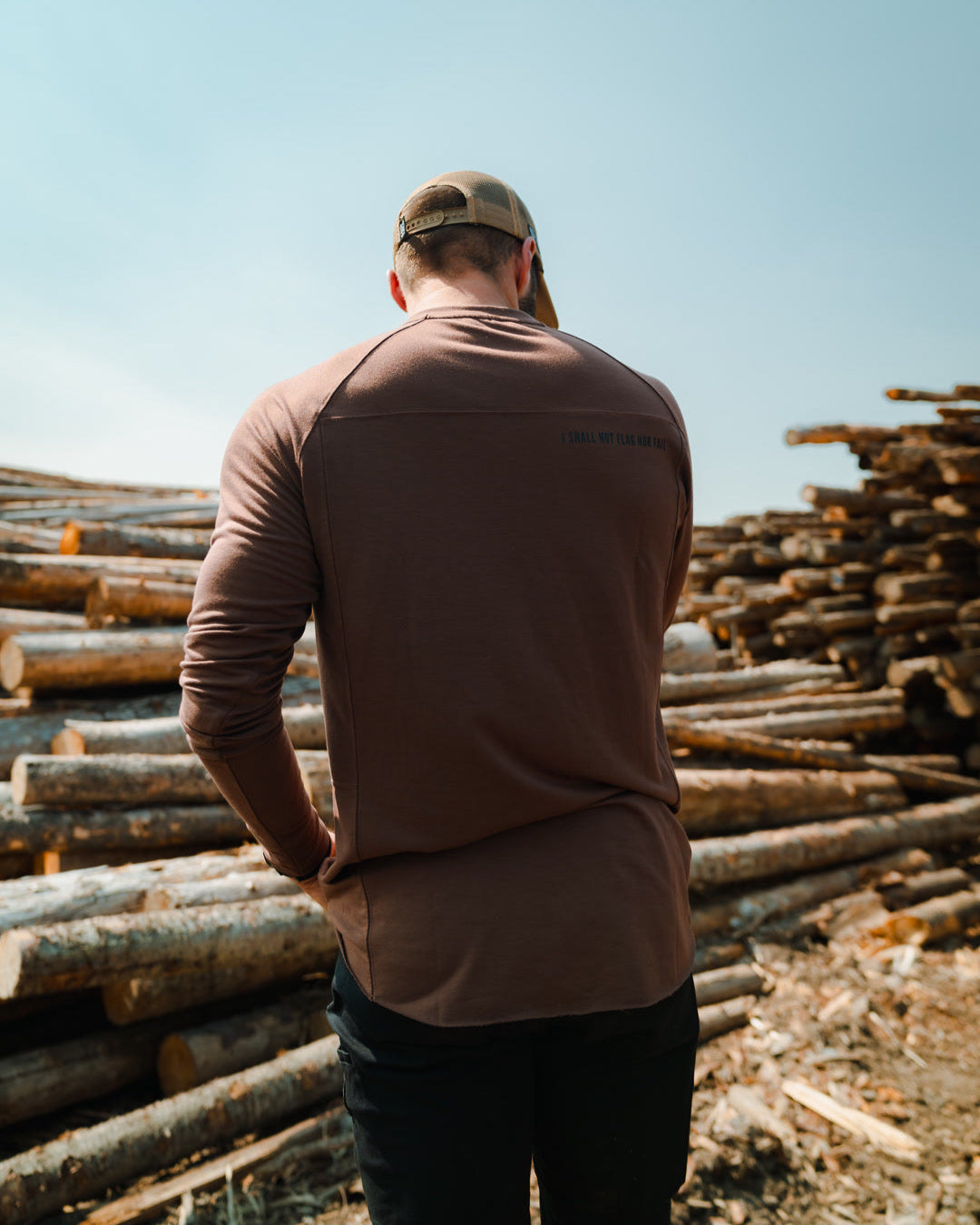 Man in a brown shirt and cap standing among stacks of logs with a clear blue sky.