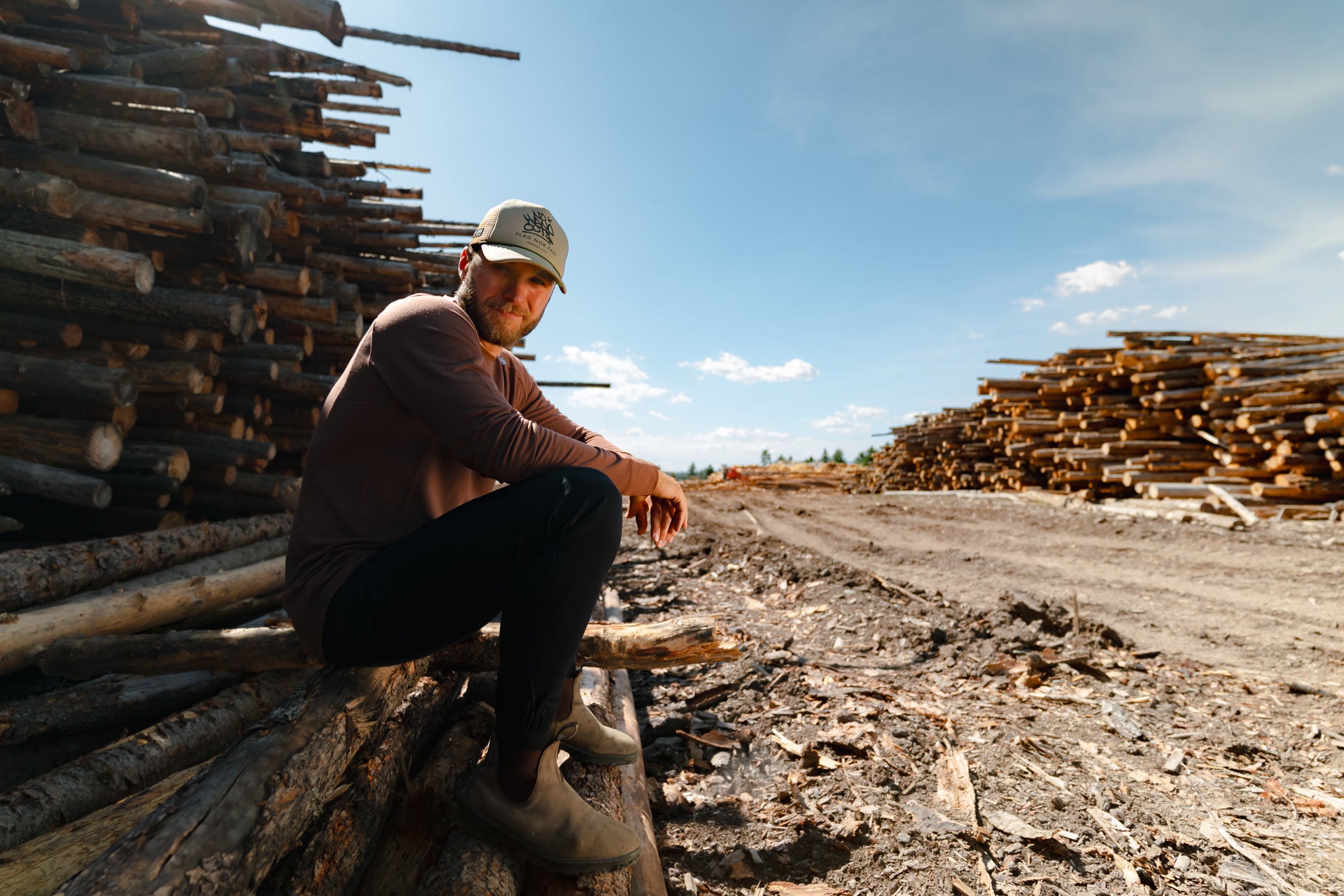 Man sitting on a stack of logs with more logs and clear sky in the background