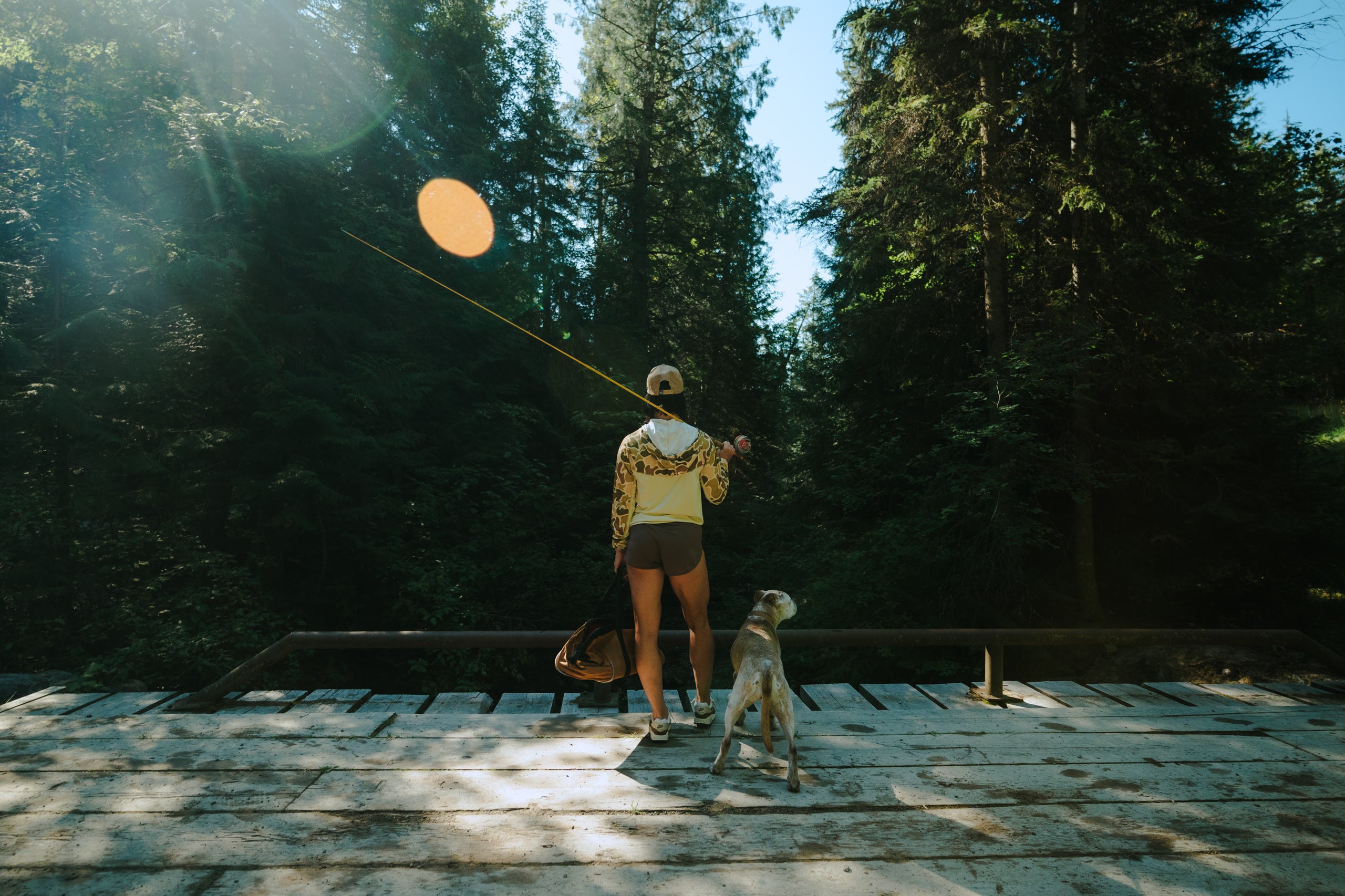 Dana and Kaiya on a wooden bridge with trees in the background overlooking a creek holding a fishing rod and tool bag