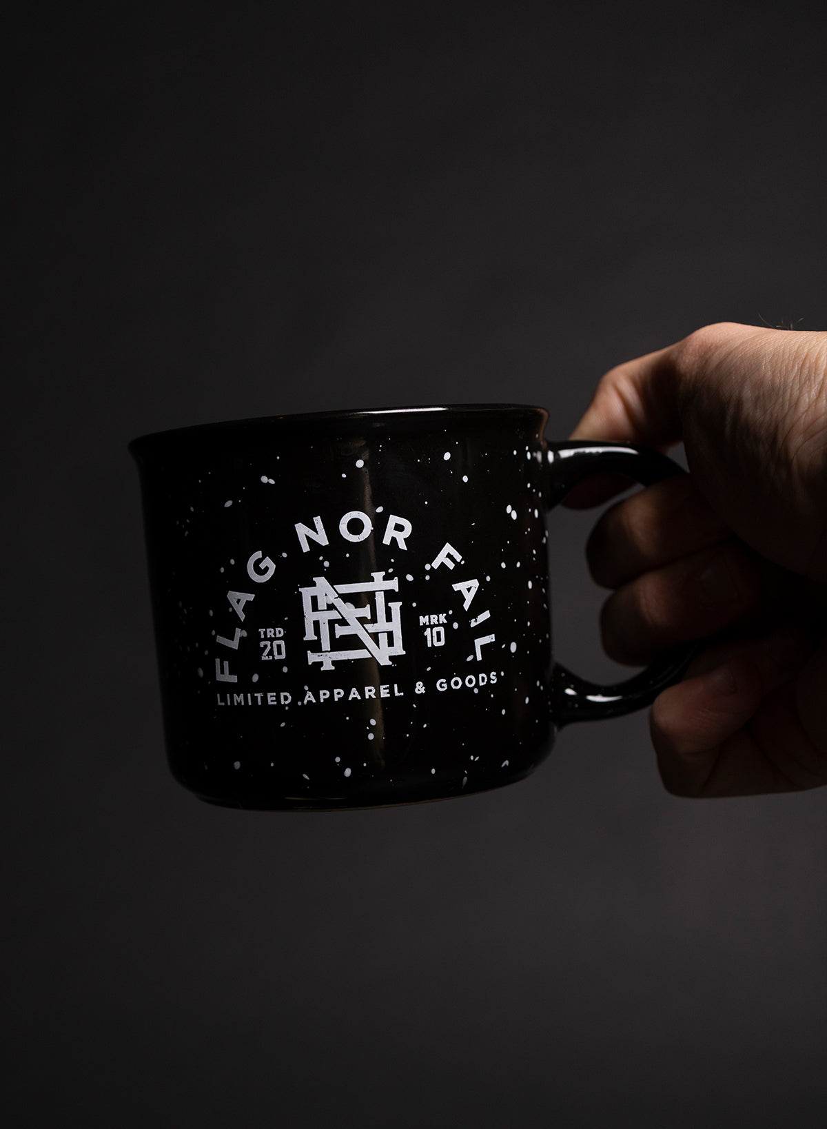 Hand holding a black ceramic mug with white speckles. Featuring a white Flag nor Fail logo.